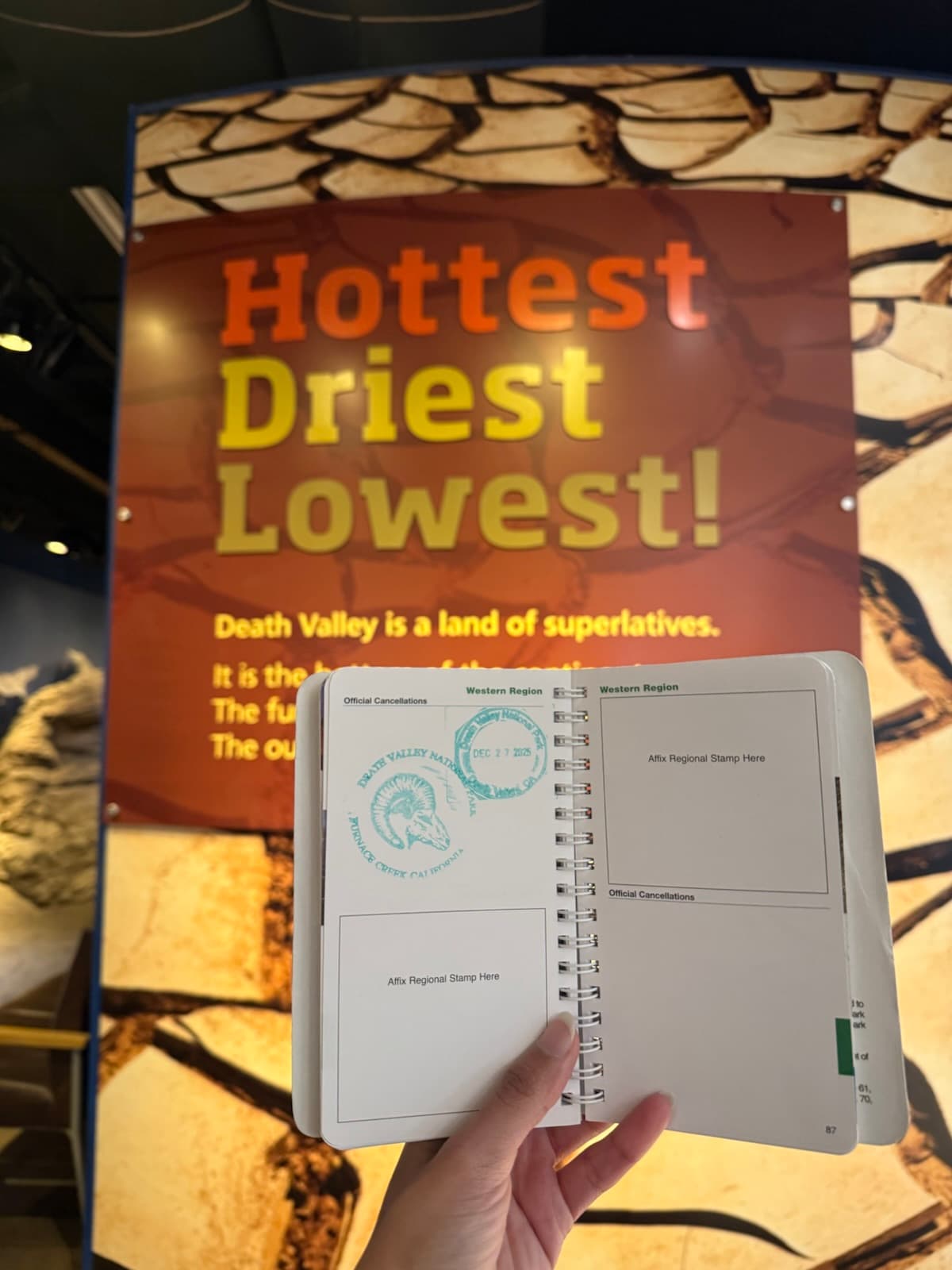 A national parks passport stamped at Death Valley, held up in front of the visitor centre's sign